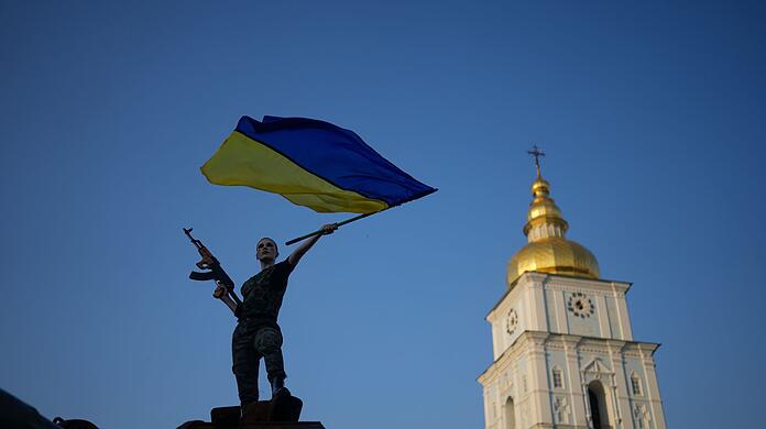 Eine Frau schwenkt die ukrainische Flagge auf einem zerstörten russischen Panzer in Kiew. Die Sonne scheint, aber es herrschen Traurigkeit und grimmige Entschlossenheit.