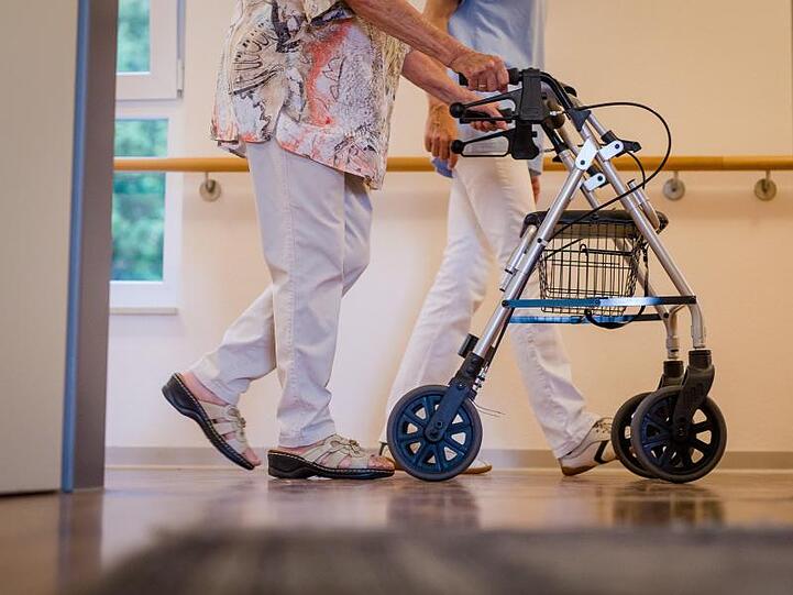 Eine Pflegekraft geht in einem Pflegeheim mit einer älteren Frau über einen Korridor. Foto: Christoph Schmidt/dpa/Archivbild