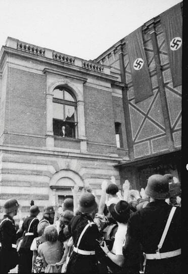 Hitler grüßt die Volksmenge von einem Fenster des Königsbaues, die Fassade des Festspielhauses ist mit Hakenkreuzfahnen geschmückt.