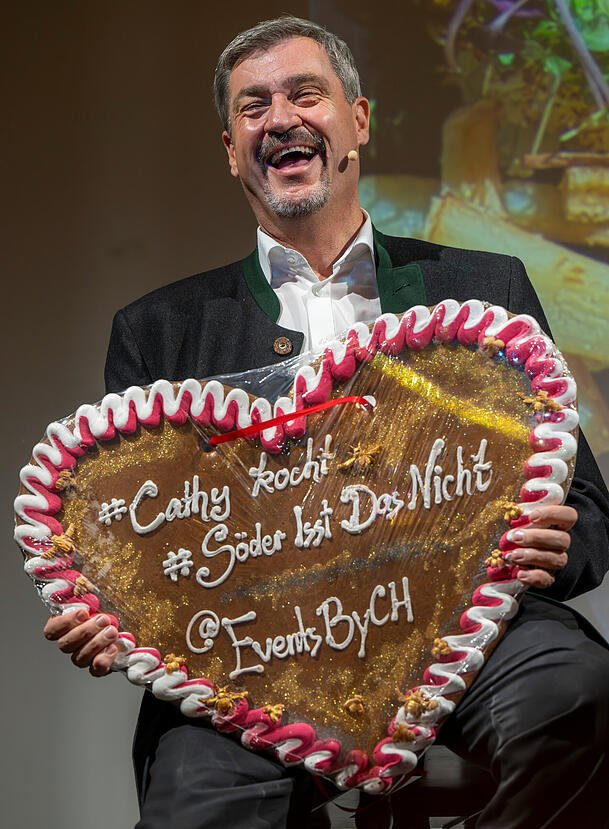 Feixend hält der Ministerpräsident von Bayern ein Lebkuchenherz mit der Aufschrift "#Cathy kocht - #Söder isst Das Nicht" in die Kamera. Feixend hält der Ministerpräsident von Bayern ein Lebkuchenherz mit der Aufschrift "#Cathy kocht - #Söder isst Das Nicht" in die Kamera.