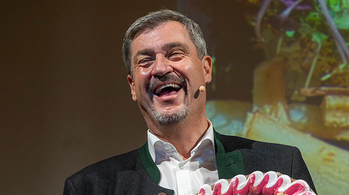Feixend hält der Ministerpräsident von Bayern ein Lebkuchenherz mit der Aufschrift "#Cathy kocht - #Söder isst Das Nicht" in die Kamera.