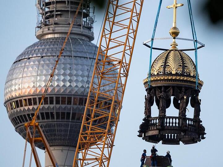 Ein Kran hebt am Neubau des Berliner Schloss die Kuppelspitze mit Engelsfiguren und einem Kreuz auf die Kuppel.