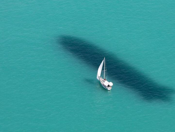 Ein Segelboot ist auf dem Bodensee unterwegs, während neben dem Boot der Schatten eines Zeppelin NT im Wasser zu sehen ist.