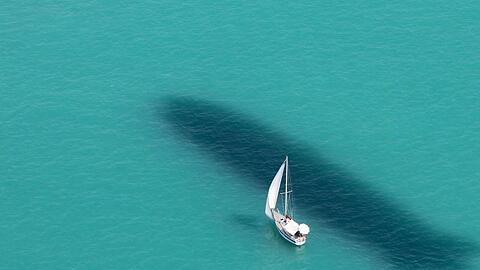 Ein Segelboot ist auf dem Bodensee unterwegs, während neben dem Boot der Schatten eines Zeppelin NT im Wasser zu sehen ist.