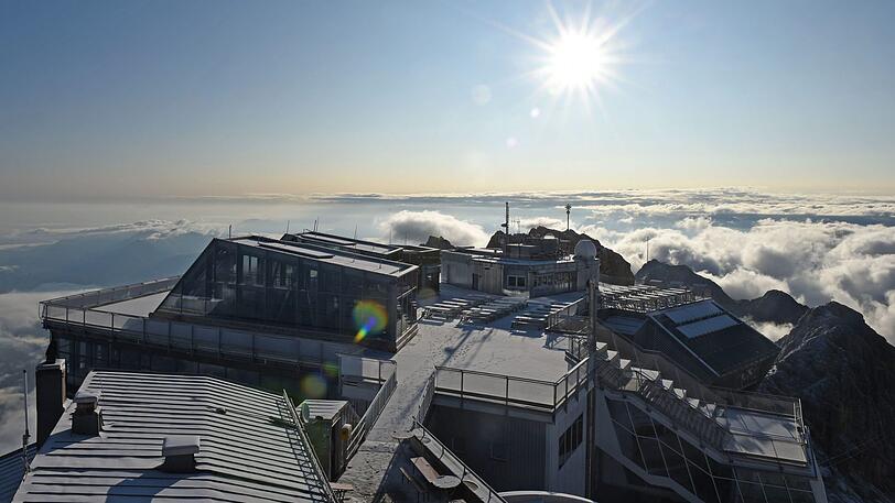 Vom Turm der Wetterstation der Zugspitze ist das Gipfelkreuz, das Dach des Münchner Hauses und die Bergstation der Seilbahn vor einem Wolkenteppich zu sehen.