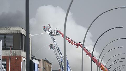 Von Drehleitern aus spritzen Feuerwehrleute Wasser auf brennende und stark rauchende Lagerhallen.