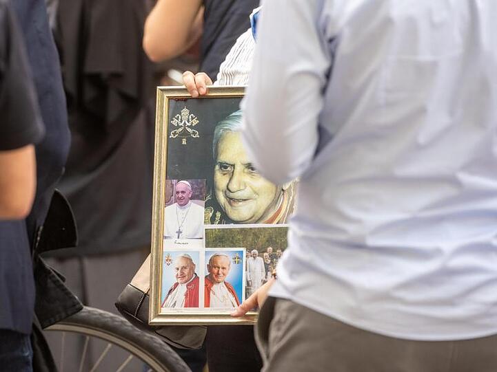 Eine Frau wartet mit einem Bild von dem emeritierte Papst Benedikt XVI. vor dem Haus von Georg Ratzinger. Foto: Armin Weigel/dpa/Archivbild