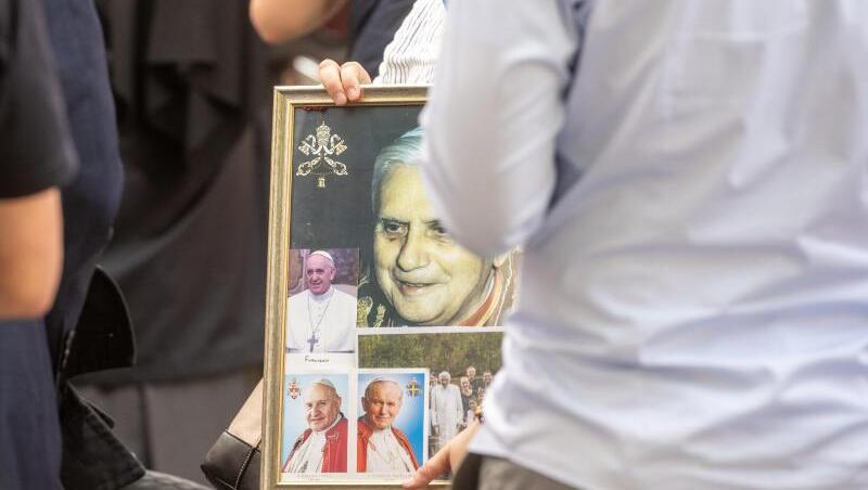 Eine Frau wartet mit einem Bild von dem emeritierte Papst Benedikt XVI. vor dem Haus von Georg Ratzinger. Foto: Armin Weigel/dpa/Archivbild Eine Frau wartet mit einem Bild von dem emeritierte Papst Benedikt XVI. vor dem Haus von Georg Ratzinger. Foto: Armin Weigel/dpa/Archivbild