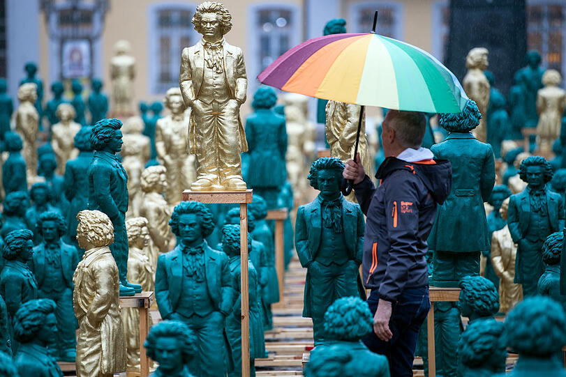 Ein Mann mit einem Schirm geht an den l&auml;chelnden Beethoven-Statuen, einem Kunstwerk des Konzeptk&uuml;nstlers und Bildhauers Ottmar H&ouml;rl, auf dem M&uuml;nsterplatz vorbei (2019).