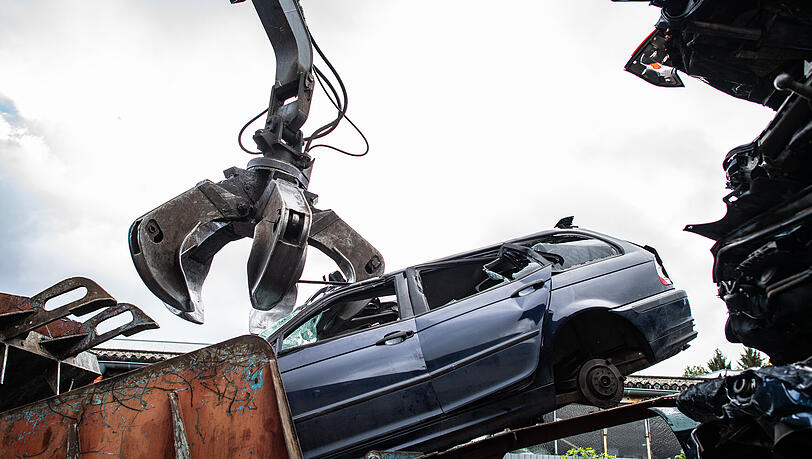 Ein Autowrack wird bei einem Recyclingbetrieb, mit dem Greifarm eines Baggers in die Schrottpresse verfrachtet. (Symbolbild)
