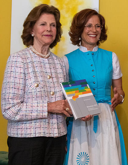 Silvia (l) bekommt im "Bayerischer Hof" von Tatjana Eckerlein, die ein original Hostessen-Dirndl der olympischen Spiele 1972 trägt, ein Buch über diese Spiele.