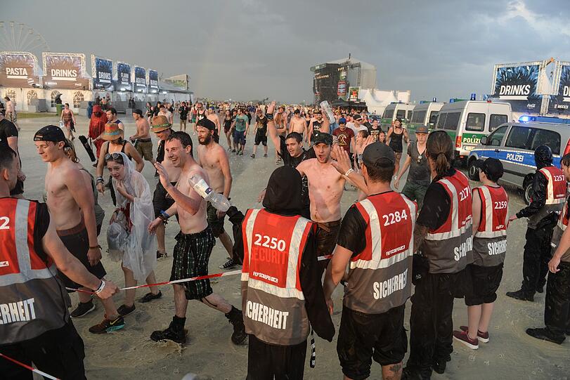 Festivalbesucher verlassen wegen einer Unwetterwarnung bei Regen das Festivalgelände, während Sicherheitskräfte das Gelände absperren.