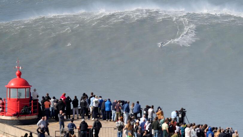 Menschen am Leuchtturm bei Nazaré beobachten einen Surfer am Praia do Norte, der eine Riesenwelle surft.
