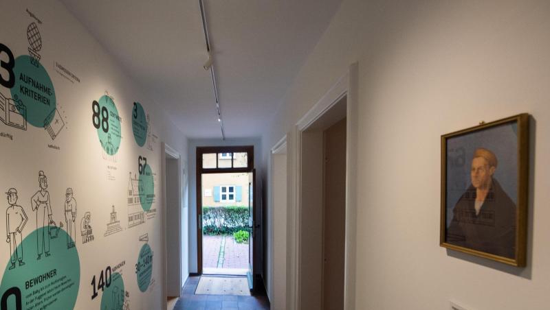 In der Fuggerei hängt ein Porträt von Jakob Fugger gegenüber einer Wand, auf der Zahlen und Fakten aufgemalt sind. Foto: Stefan Puchner In der Fuggerei hängt ein Porträt von Jakob Fugger gegenüber einer Wand, auf der Zahlen und Fakten aufgemalt sind. Foto: Stefan Puchner