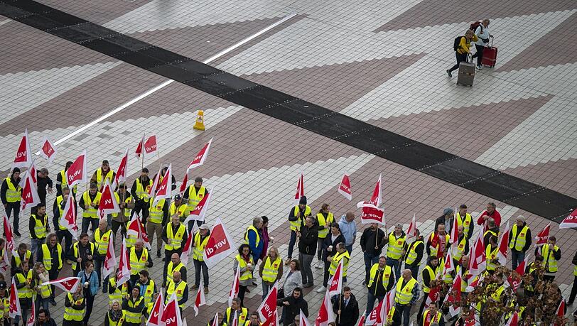Zwei Flugpassagiere mit Rollkoffern laufen an Streikenden im Zentralbereich des Flughafens München vorbei, die an einer Protestaktion teilnehmen.