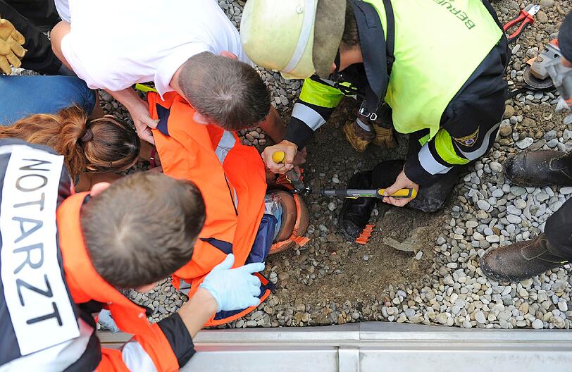 Mit vereinten Kräften wird das Kind gerettet: Ein Feuerwehrmann schaufelt das Rohr frei, einer hält eine Zange zum Aufschneiden in der Hand, wieder andere beruhigen den Kleinen. Auch die Mutter spricht beruhigend auf ihren Sohn ein.