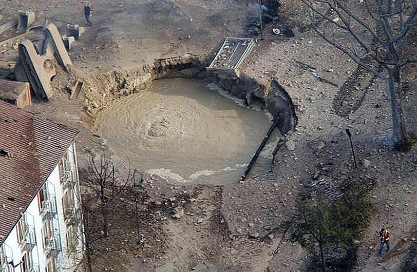 Ein durch geborstene Leitungen mit Wasser gefüllter Exploionskrater klafft zwischen einem Wohnhaus und einer zerstörten Straße. Neben dem Krater ragt der umgestürzte Bagger hervor. Ersten Ermittlungen zufolge war es bei Arbeiten an einer Gasleitung zu dem Unglück gekommen.