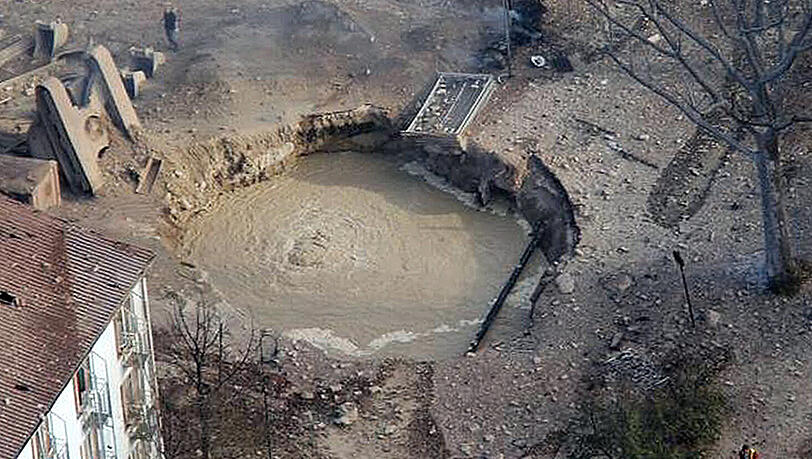 Ein durch geborstene Leitungen mit Wasser gefüllter Exploionskrater klafft zwischen einem Wohnhaus und einer zerstörten Straße. Neben dem Krater ragt der umgestürzte Bagger hervor. Ersten Ermittlungen zufolge war es bei Arbeiten an einer Gasleitung zu dem Unglück gekommen. Ein durch geborstene Leitungen mit Wasser gefüllter Exploionskrater klafft zwischen einem Wohnhaus und einer zerstörten Straße. Neben dem Krater ragt der umgestürzte Bagger hervor. Ersten Ermittlungen zufolge war es bei Arbeiten an einer Gasleitung zu dem Unglück gekommen.