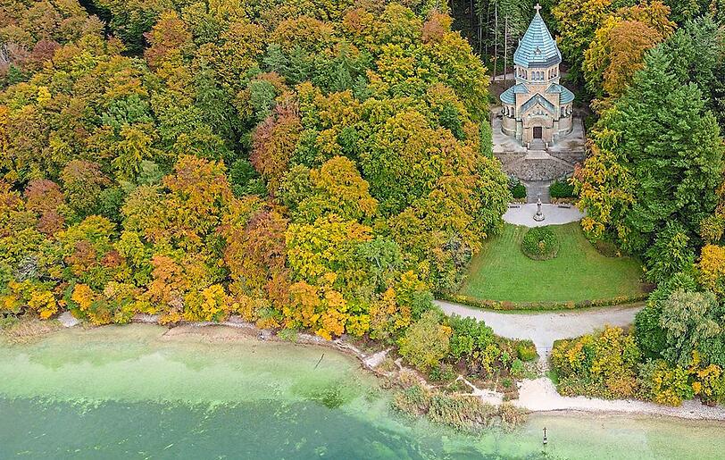 Die Votivkapelle zum Gedenken an den Kini in Berg, umgeben von Natur. Am Ufer davor ist das Kreuz für Ludwig II. zu erkennen.