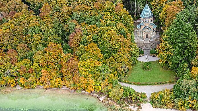 Die Votivkapelle zum Gedenken an den Kini in Berg, umgeben von Natur. Am Ufer davor ist das Kreuz für Ludwig II. zu erkennen.