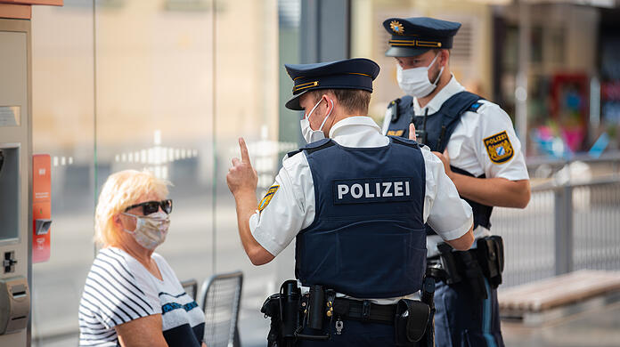 Zwei Polizisten nehmen an einer Tram-Haltestelle von einer Frau nach einem Verstoß gegen die Maskenpflicht im öffentlichen Nahverkehr die Personalien auf.