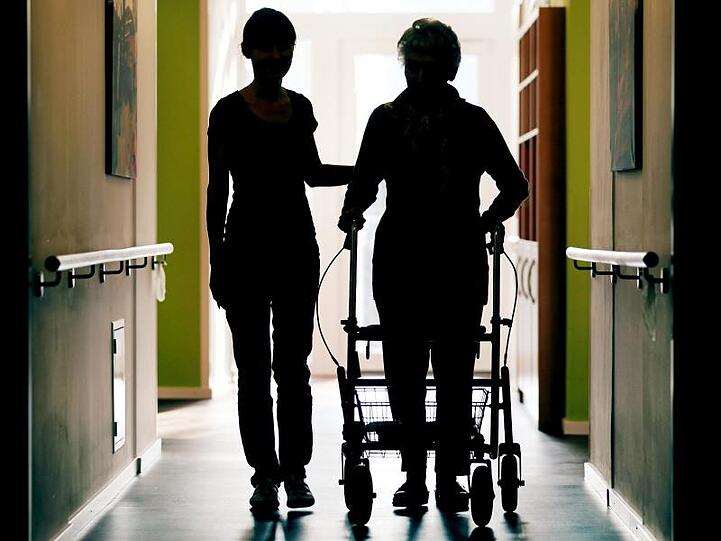 Eine Pflegekraft begleitet die Bewohnerin eines Altenheims mit Rollator beim Gang durch den Flur. Foto: Oliver Berg/dpa/Archivbild