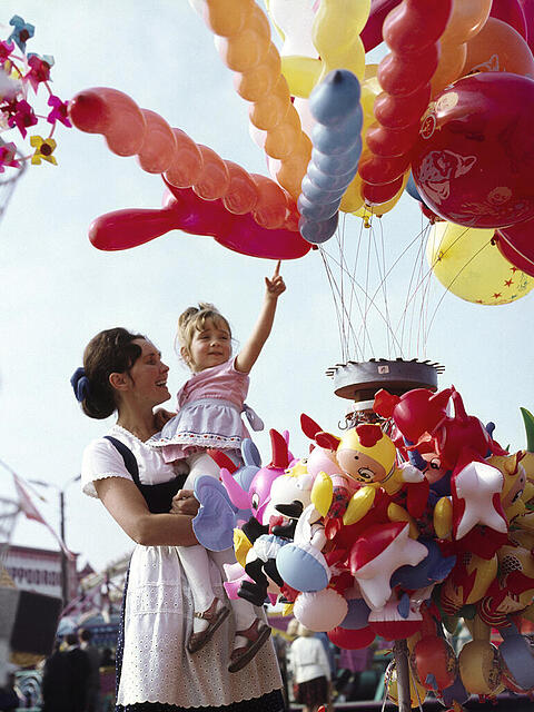 Am Fr&uuml;hlingsfest in den 60er Jahren: Eine fr&ouml;hliche Mutter h&auml;lt ihre Tochter auf dem Arm, die einen Luftballon erhaschen m&ouml;chte. Ob sie einen bekommen hat?