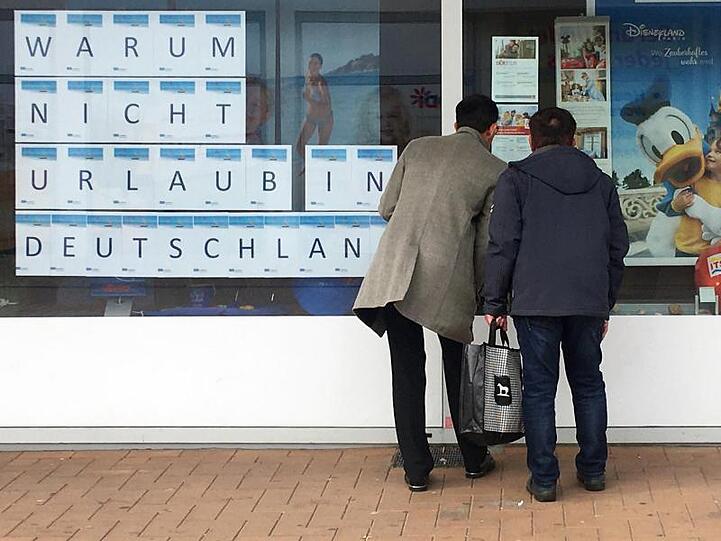 Zwei Männer schauen in das Schaufenster eines Reisebüros, an dem mit großen Buchstaben "Warum nicht Urlaub in Deutschland" zu lesen ist.