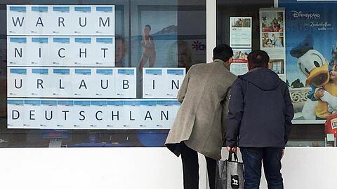 Zwei Männer schauen in das Schaufenster eines Reisebüros, an dem mit großen Buchstaben "Warum nicht Urlaub in Deutschland" zu lesen ist.