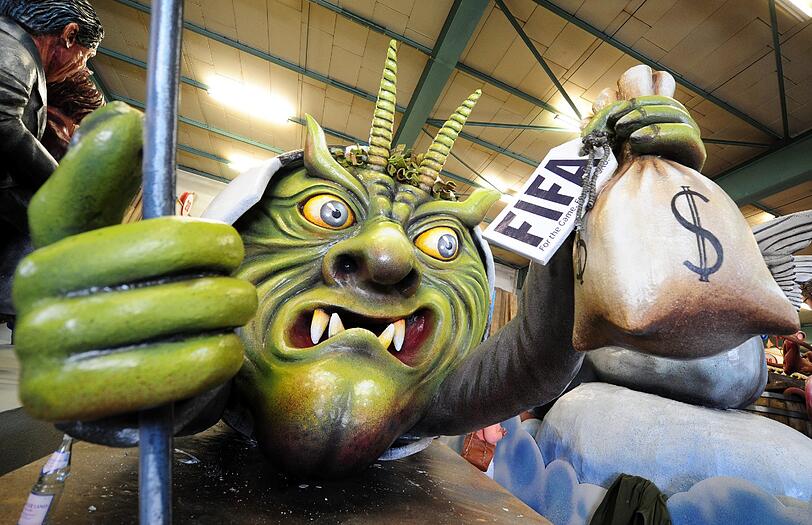 Der Mainzer Motivwagen "Die halbe Wahrheit" zeigt eine grüne Teufelsfigur, die aus der Rückseite eines Fußballs hervorkommt und einem Geldsack hält. Das Motiv symbolisiert den Fußballverband FIFA.