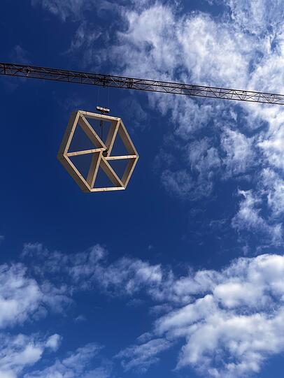 Ein Sechseck schwebt vor weiß-blauem Himmel zur  Baustelle und wird dort montiert.