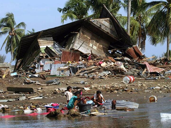 Frauen und Kinder, die den Tsunami in der Nähe von Banda Aceh überlebt haben, hocken vor einem zerstörten Haus an einer Wasserstelle.