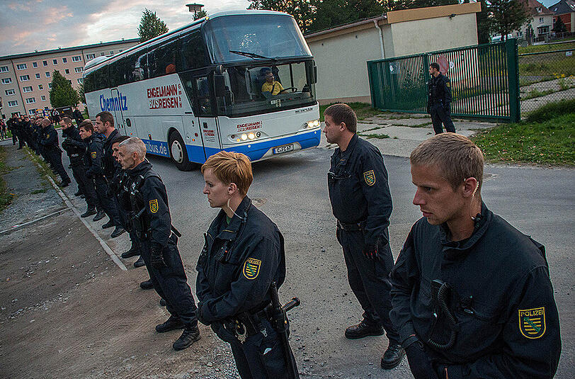 Polizisten stehen am 18.09.2015 in Bischofswerda vor einem Bus, der Flüchtlinge in eine Notunterkunft bringt. Mehr als 50 Menschen versammelten sich am Freitagabend vor dem früheren Bekleidungsbetrieb und skandierten fremdenfeindliche Parolen.