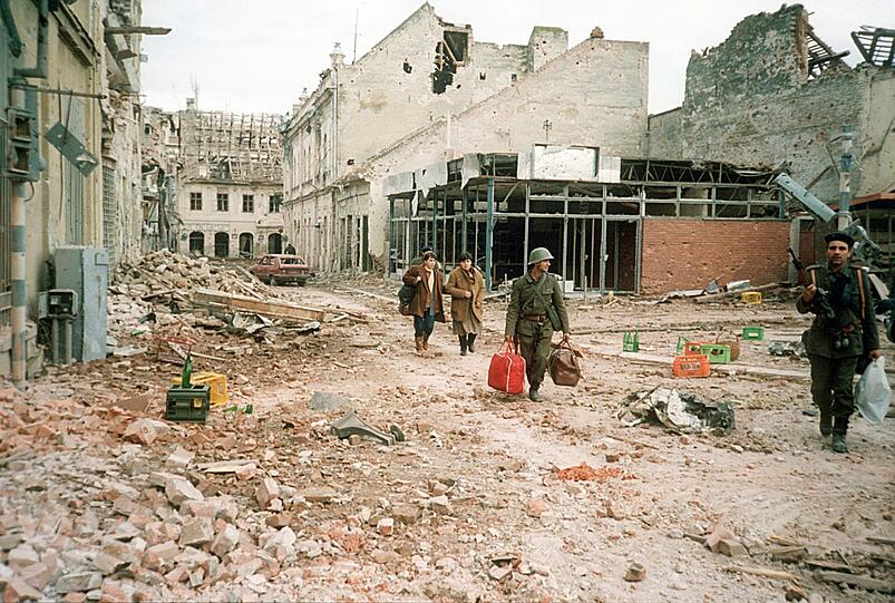Das Archivbild zeigt Menschen, die am 19.11.1991 einen zerstörten Straßenzug in der Stadt Vukovar entlang gehen. In der Ausstellung zeigen Künstler einen Blick auf ihre Kindheit in Jugoslawien.