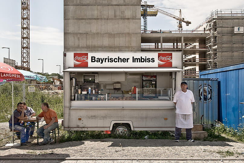 Im Juli 2000 ist Spatenstich fürs Mega-Neubaugebiet Parkstadt Schwabing - ab da sind auf dem Areal noch über Jahre Kräne zu sehen. Klar braucht es für die vielen Arbeiter vor Ort vor allem drei Dinge: Kaffee, Leberkässemmeln und Franziskaner-Weißbier. Die gibt es bei diesem Imbisswagen, der orthografisch nicht ganz sauber "Bayrischer Imbiss" heißt. Ein recht gemütliches Pausenfleckerl mitten unter den Rohbauten ist das - wie man den drei Bauarbeitern links unter den Sonnenschirmen recht schön ansehen kann.
