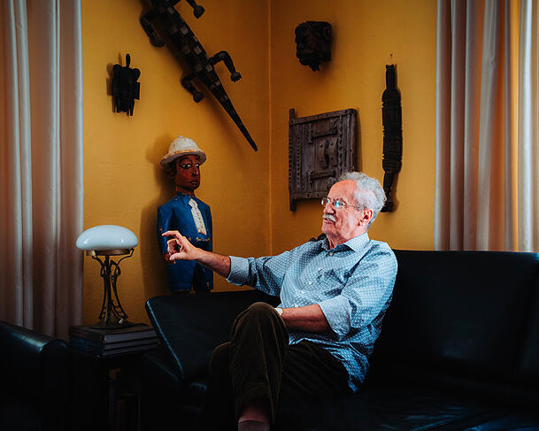Ude sitzt in seinem Wohnzimmer auf einer Ledercouch, dahinter an der Wand sind Figuren von Afrikareisen.