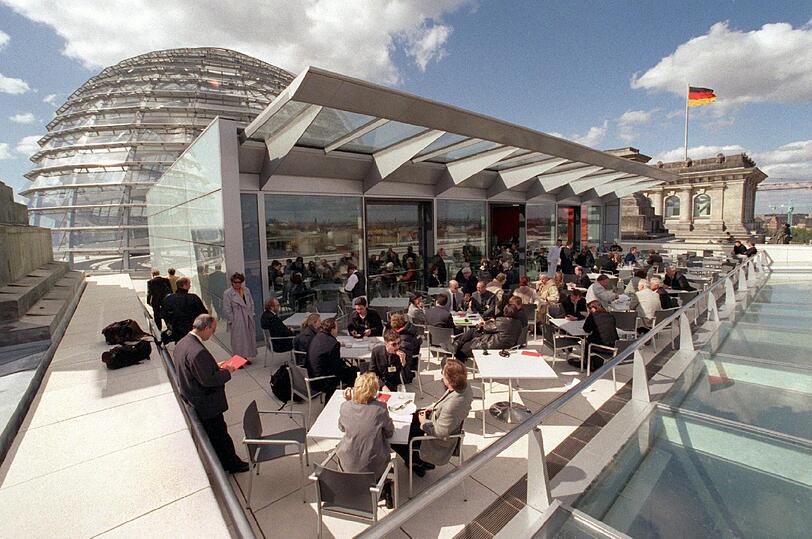 Im Sonnenlicht sitzen Abgeordnete und Besucher auf der Terrasse des Berliner Reichstagsgebäudes. Im Hintergrund weht über einem der Türme des historischen Bauwerks die deutsche Fahne.