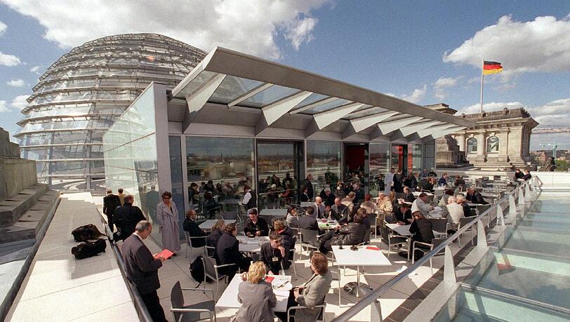 Im Sonnenlicht sitzen Abgeordnete und Besucher auf der Terrasse des Berliner Reichstagsgebäudes. Im Hintergrund weht über einem der Türme des historischen Bauwerks die deutsche Fahne. Im Sonnenlicht sitzen Abgeordnete und Besucher auf der Terrasse des Berliner Reichstagsgebäudes. Im Hintergrund weht über einem der Türme des historischen Bauwerks die deutsche Fahne.