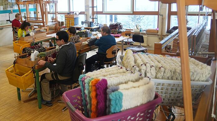 Blinde und sehbehinderte Kollegen arbeiten in ihrem Handwerk in der SWW-Weberei. Im Vordergrund sind Sitzkissen für die Münchner Kammerspiele zu sehen.