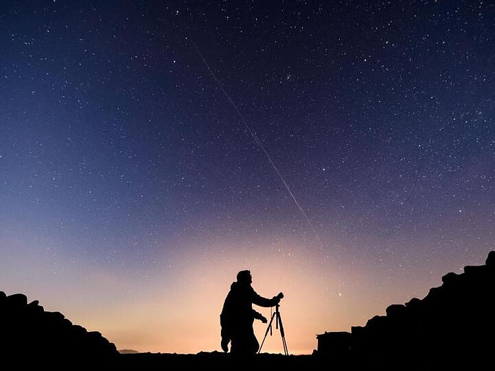 Ein Fotograf steht vor einem Nachthimmel, an dem die Bewegung von Starlink-Satelliten als leuchtende Spur (Bildmitte) sichtbar wird.