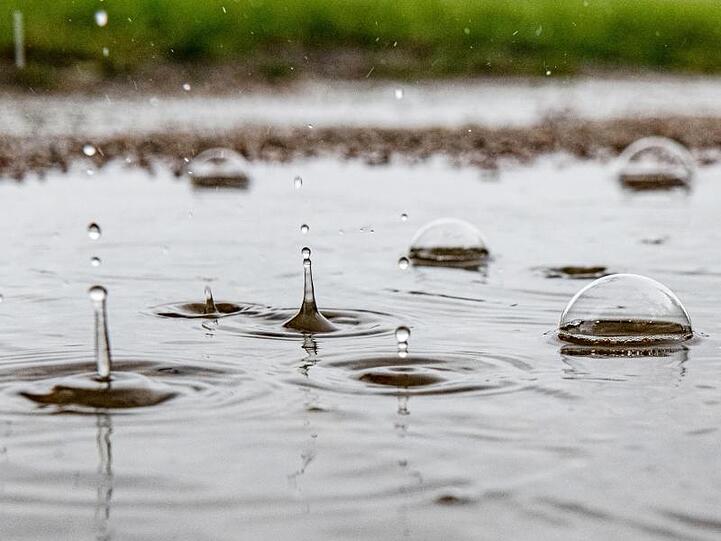 Regentropfen fallen in eine Pfütze und erzeugen durch den Aufprall kunstvolle Gebilde. Foto: Paul Zinken/dpa/Symbolbild
