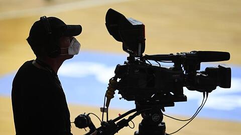 Ein Kameramann steht mit Mundschutz bei einem Basketballspiel an seiner TV-Kamera.