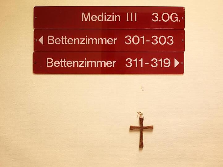 Ein kleines Holzkreuz hängt unter Hinweisschildern auf einer Station in einem kirchlichen Krankenhaus.