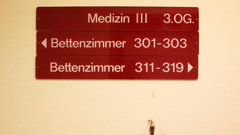 Ein kleines Holzkreuz hängt unter Hinweisschildern auf einer Station in einem kirchlichen Krankenhaus. Ein kleines Holzkreuz hängt unter Hinweisschildern auf einer Station in einem kirchlichen Krankenhaus.