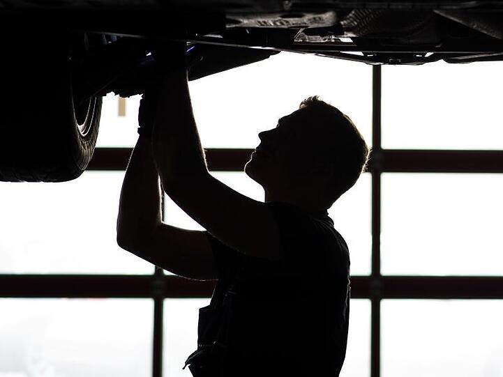 Ein Mechaniker arbeitet in einer KfZ-Werkstatt an der Unterseite eines Autos, dass auf der Hebebühne steht. Foto: David Ebener/dpa/Archivbild