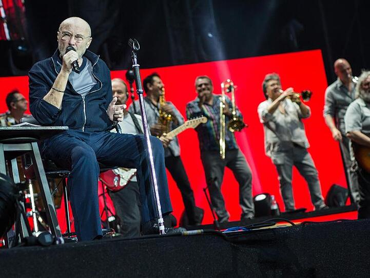 Sänger Phil Collins sitzt auf der Bühne und singt, hinter ihm stehen die Bandmitglieder.