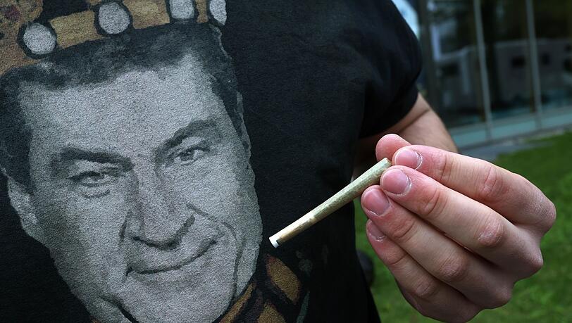 Ein Teilnehmer einer Versammlung, die unter dem Motto "Liberalitas Bavariae statt CSU-Verbotsirrtum" angemeldet wurde, hält am Gebäude der CSU-Landesleitung vor einem T-Shirt mit einem Portrait des bayerischen Ministerpräsidenten Markus Söder (CSU), einen Joint.