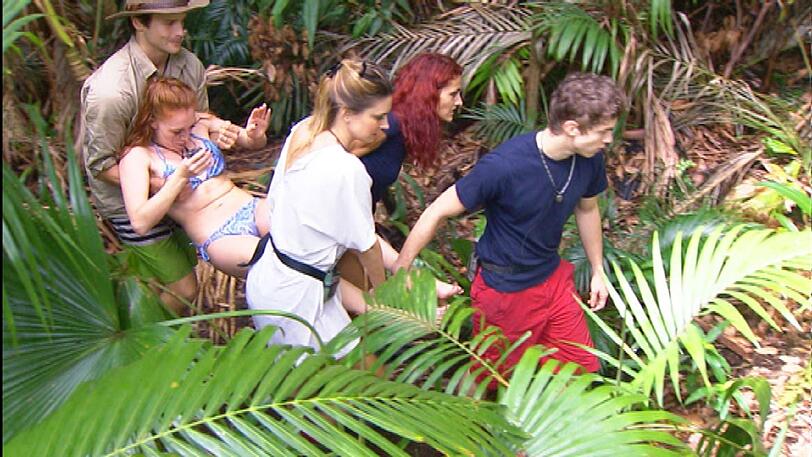 Patrick, Fiona, Claudelle und dann auch Joey tragen Georgina an Händen und Füßen zum Dschungeltelefon. Olivia: &bdquo;Lasst sie bloß nicht fallen.&ldquo; Olivia geht voran. Georgina: &bdquo;Hauptsache den Nägeln passiert nichts.&ldquo; Olivia: &bdquo;Ach Mensch, jetzt hör auf mit den Nägeln.&ldquo; Fiona: &bdquo;Die Alte &hellip;. Claudelle: &bdquo;Da war der vollgepinkelte Topf von Klaus schwerer.&ldquo;