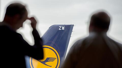 Zwei Passagiere blicken auf einem Flughafen durch ein Fenster auf eine abgestellte Lufthansa-Maschine.