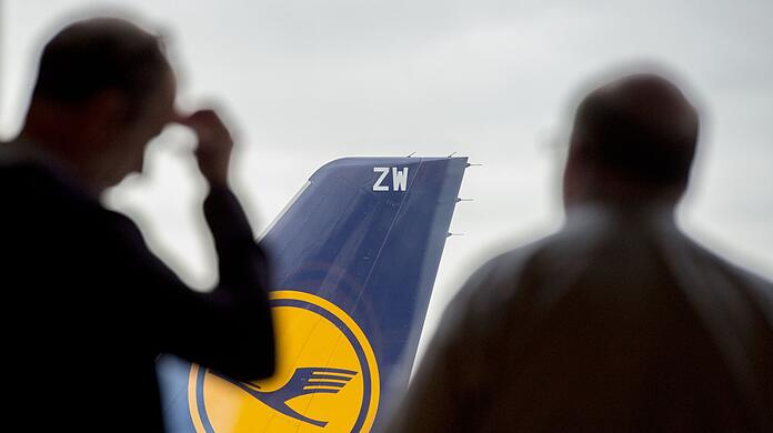 Zwei Passagiere blicken auf einem Flughafen durch ein Fenster auf eine abgestellte Lufthansa-Maschine.